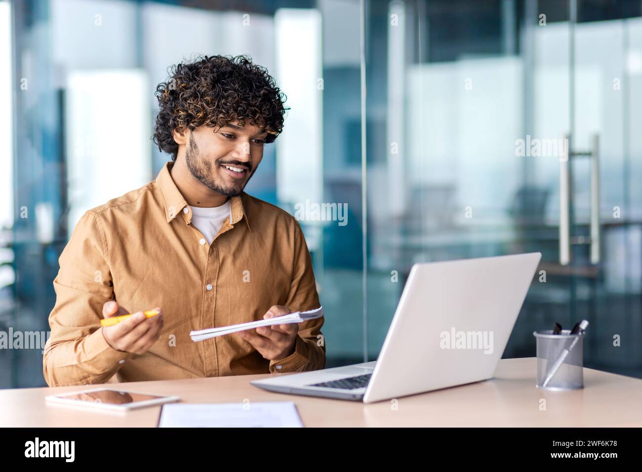 Uomo arabo attento che tiene un blocco note e una penna mentre si siede accanto al tavolo con un computer portatile in un luogo di lavoro moderno. Freelance positivi che scrivono i desideri e le idee dei clienti sulla realizzazione dei progetti tramite videochiamata. Foto Stock