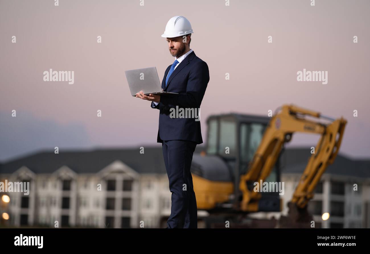 Proprietario di un'impresa edile. Uomo con tuta e cappello duro al cantiere edile. Ingegnere civile di fronte alla casa vicino all'escavatore. Arco Foto Stock