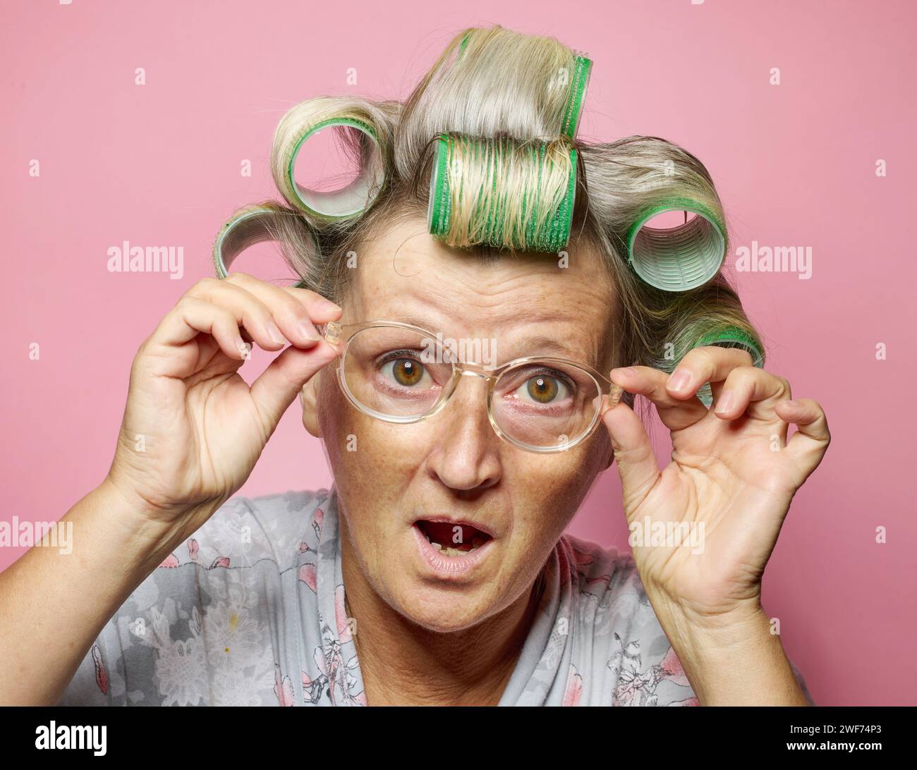 divertente, stupita, sorprende le donne anziane con rotoli di capelli e occhiali su sfondo rosa Foto Stock