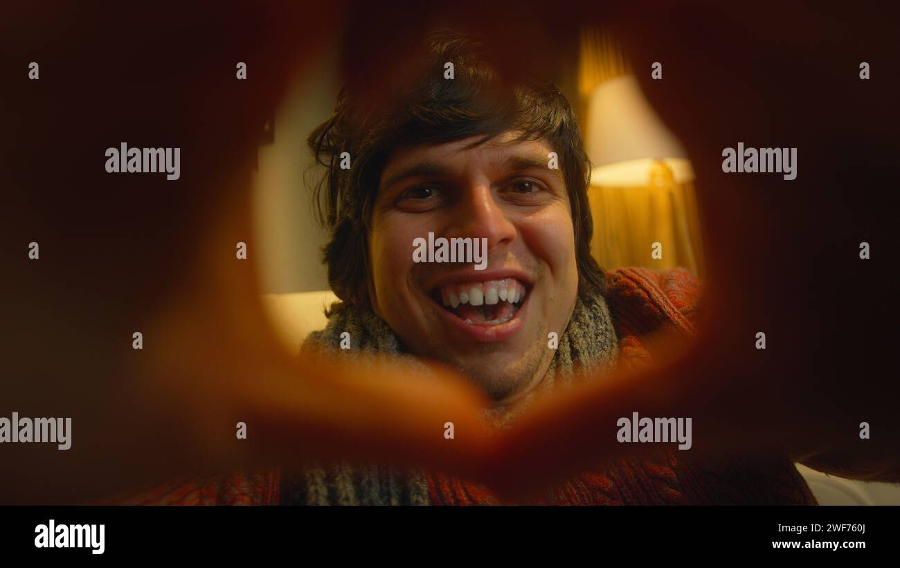 Ritratto di un giovane uomo che indossa una sciarpa che crea una forma a cuore con le dita unite che guardano la fotocamera con un sorriso tra le dita sagomate Foto Stock