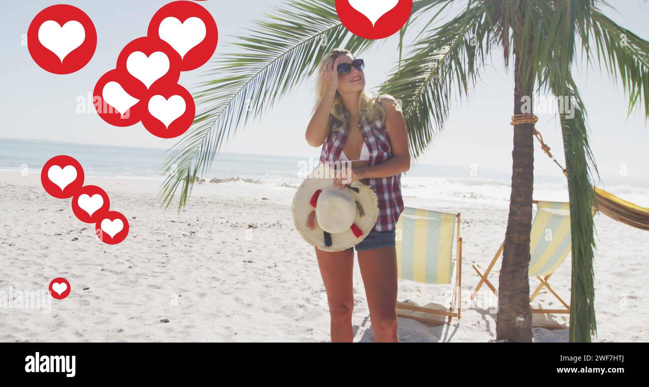 Più icone del cuore che fluttuano contro la donna caucasica con un cappello che regola i capelli sulla spiaggia Foto Stock