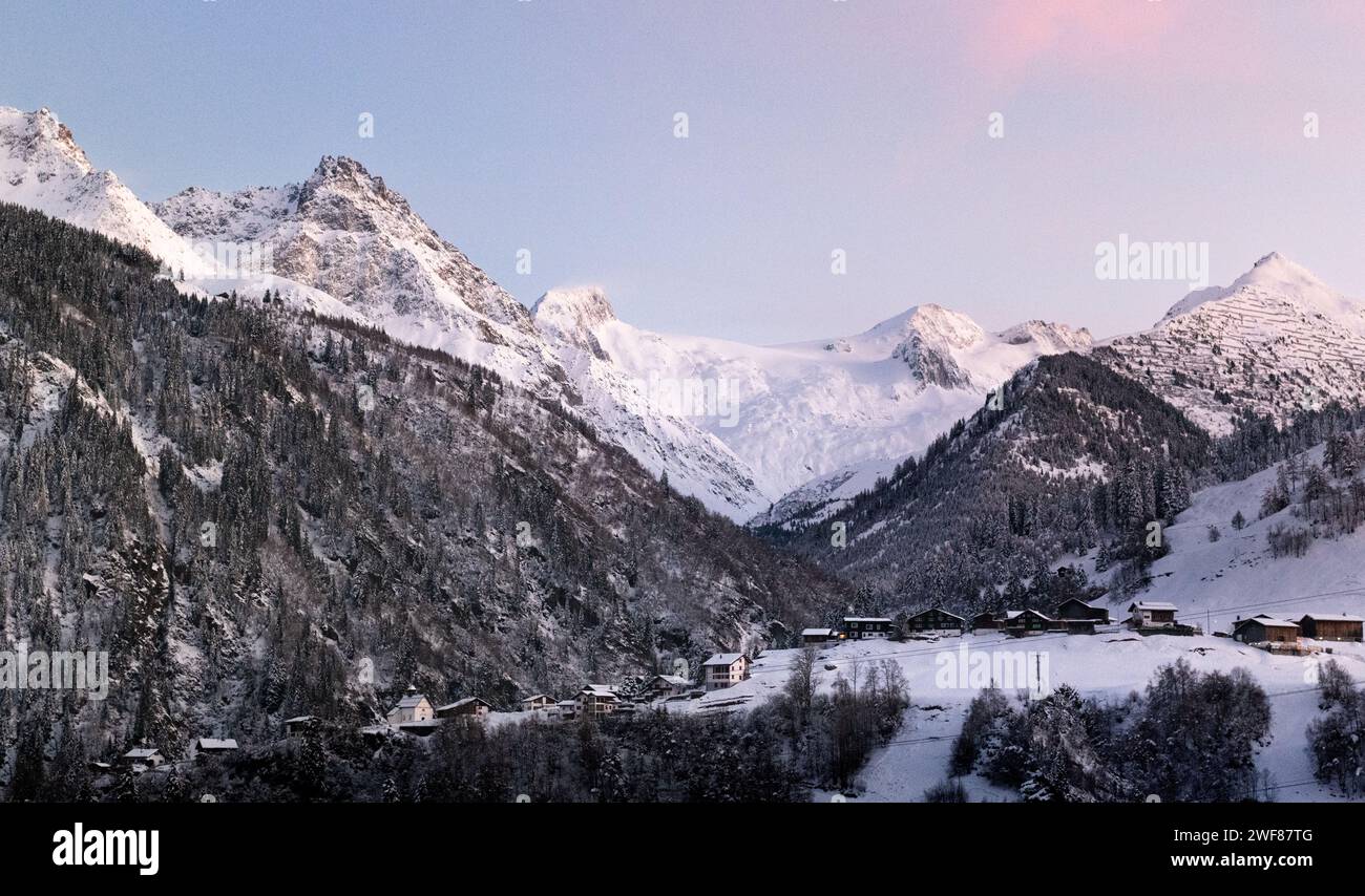 La magia invernale nelle Alpi svizzere: Un villaggio, una foresta innevata e cime dalle tonalità rosa sotto il sole serale. Un grande ghiacciaio aggiunge al pittoresco panorama. Foto Stock