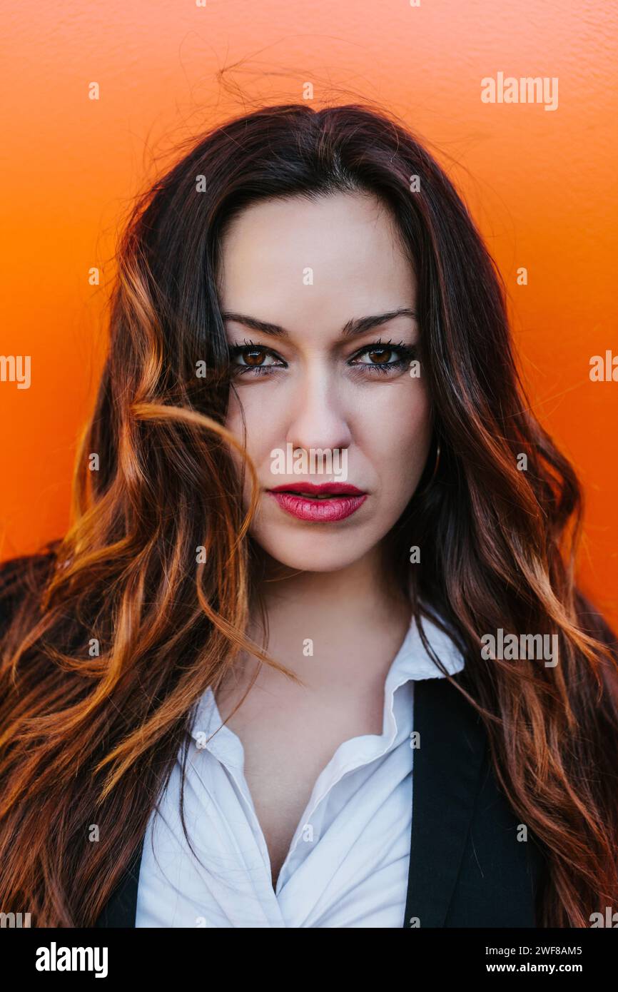 Una giovane donna in posa con camicia bianca e gilet nero regala uno sguardo penetrante con i suoi occhi intriganti, contrastati da un vivace sfondo arancione in Mad Foto Stock