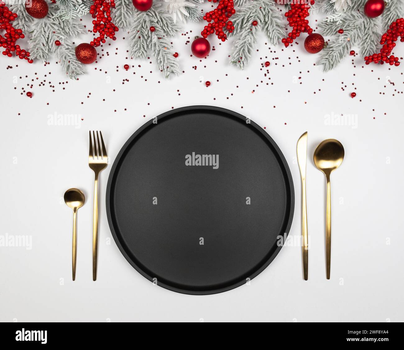 Composizione natalizia, piatto nero e posate d'oro, rami di abete, bacche rosse, bauble su sfondo bianco. Foto Stock