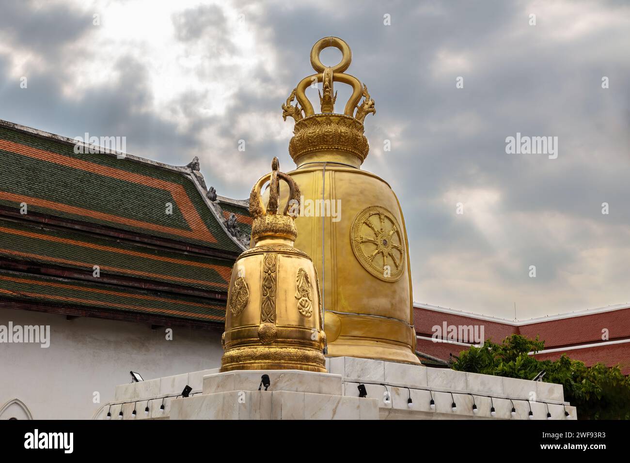 Campane dorate in mostra al Wat Rakhang (Tempio delle campane), Bangkok, Thailandia. Tetto in piastrelle colorate dietro con piastrelle arancioni e verdi. Cielo nuvoloso Foto Stock