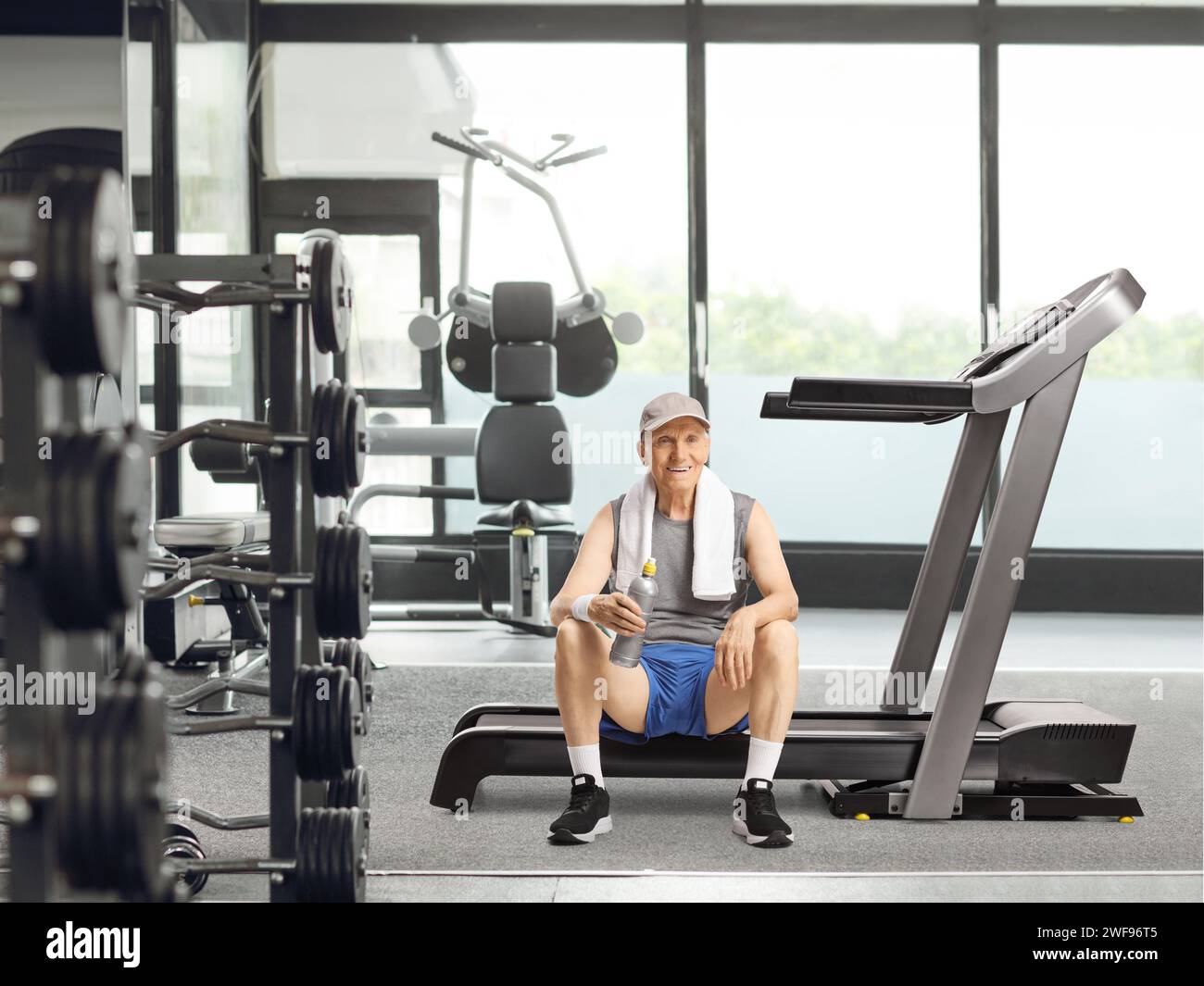 Anziano seduto su un tapis roulant in una palestra con una bottiglia d'acqua e sorridendo davanti alla macchina fotografica Foto Stock