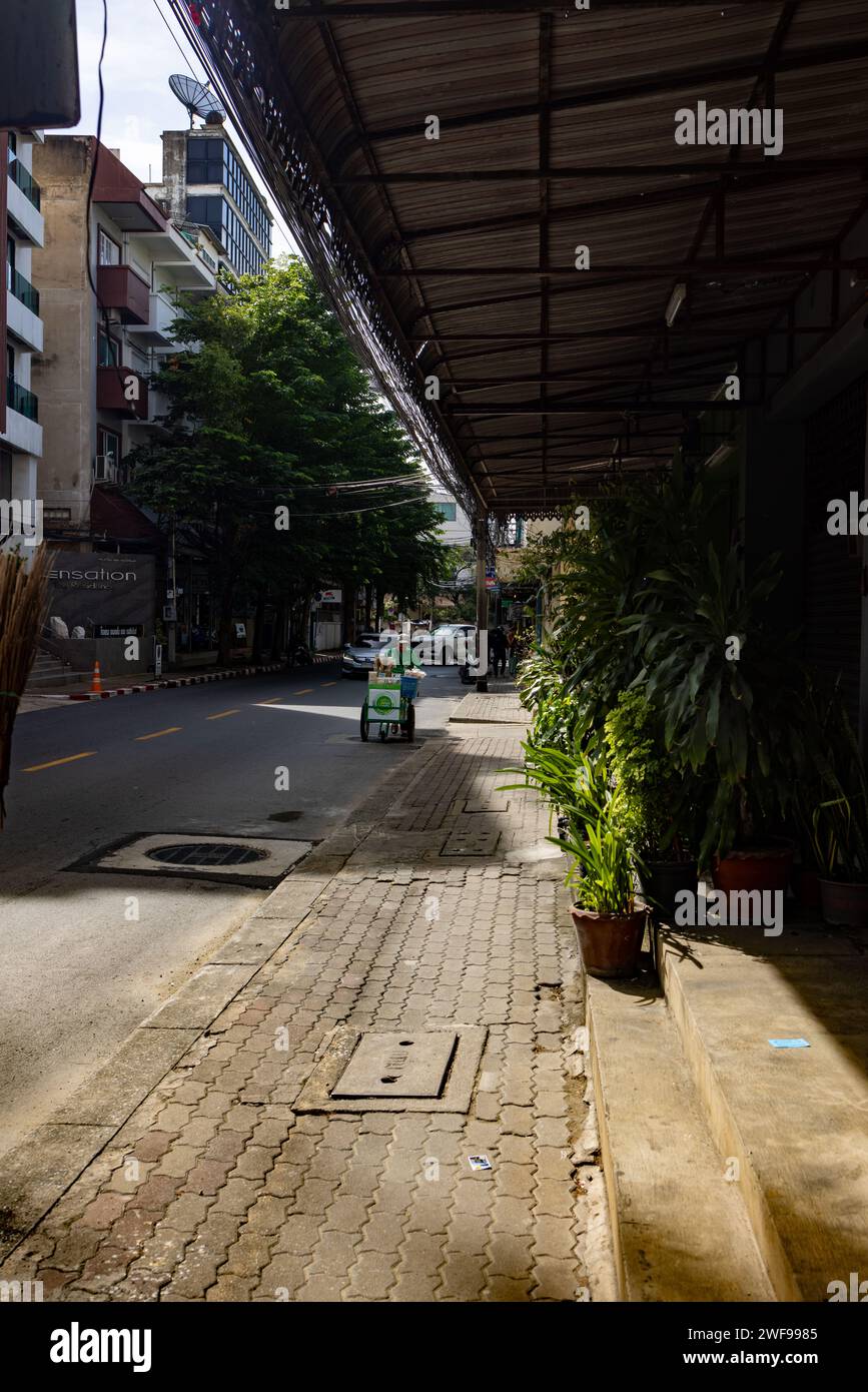 L'immagine cattura un marciapiede tranquillo e soleggiato in un'area urbana, fiancheggiata da edifici e disseminata di piante in vaso, mentre alcuni veicoli passano nel Foto Stock