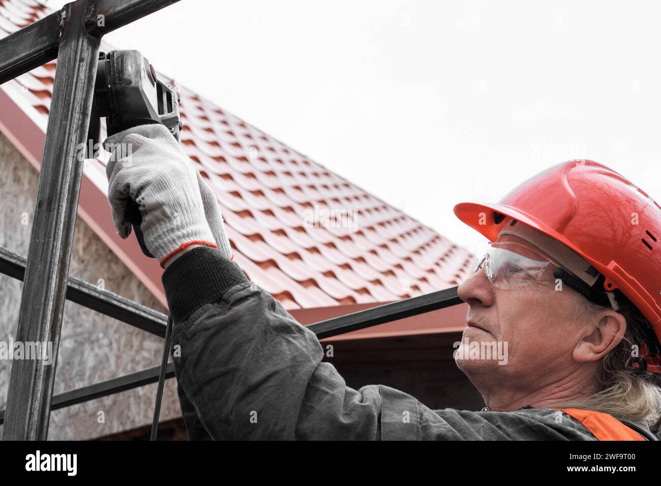 Un lavoratore che indossa un casco e occhiali di sicurezza lavora con una smerigliatrice con un tubo profilato. Installazione e costruzione. Foto Stock