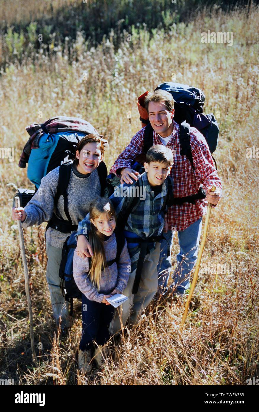 Un Ritratto di una famiglia felice con due bambini e zaini sorride alla macchina fotografica mentre si fa un'escursione in un campo erboso in una giornata di sole autunno. Foto Stock