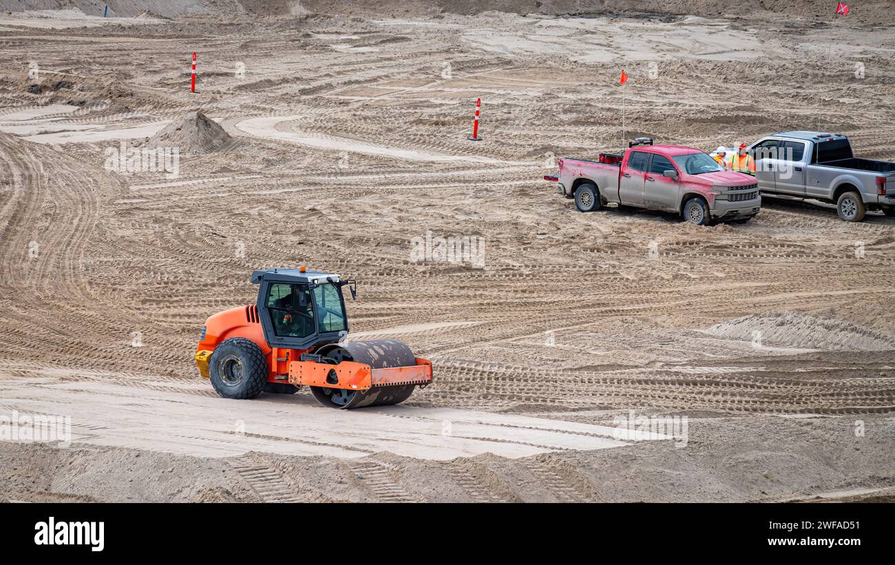 Compattatore vibrante a rulli per terreno che compatte uno spessore di sabbia in cantiere, con due raccoglitori e lavoratori Foto Stock