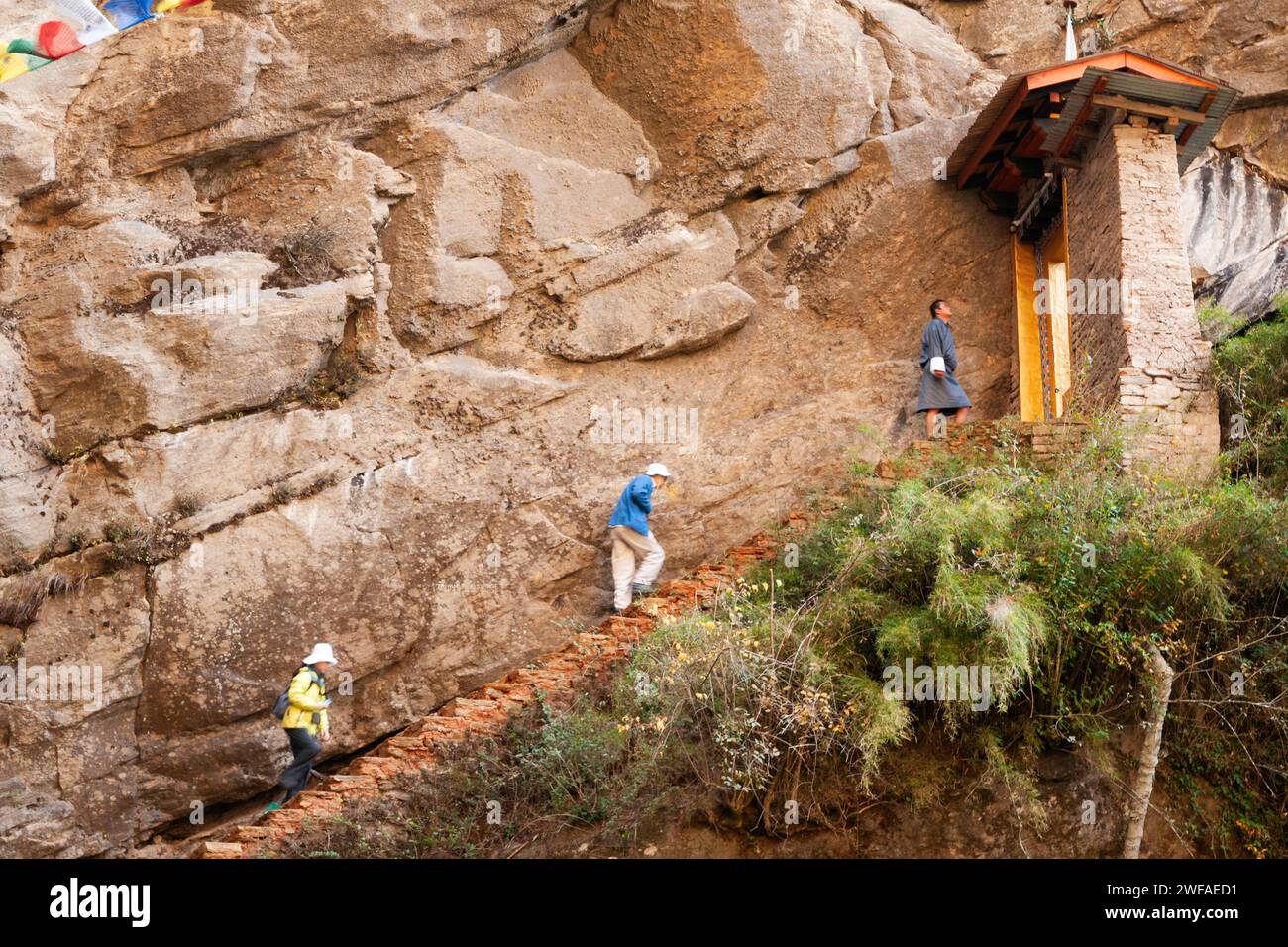 Un uomo bhutanese che indossa gho tradizionale e turisti femminili salgono le ripide scale sul sentiero roccioso del monastero Tiger's Nest, pronto a passare attraverso un giorno Foto Stock
