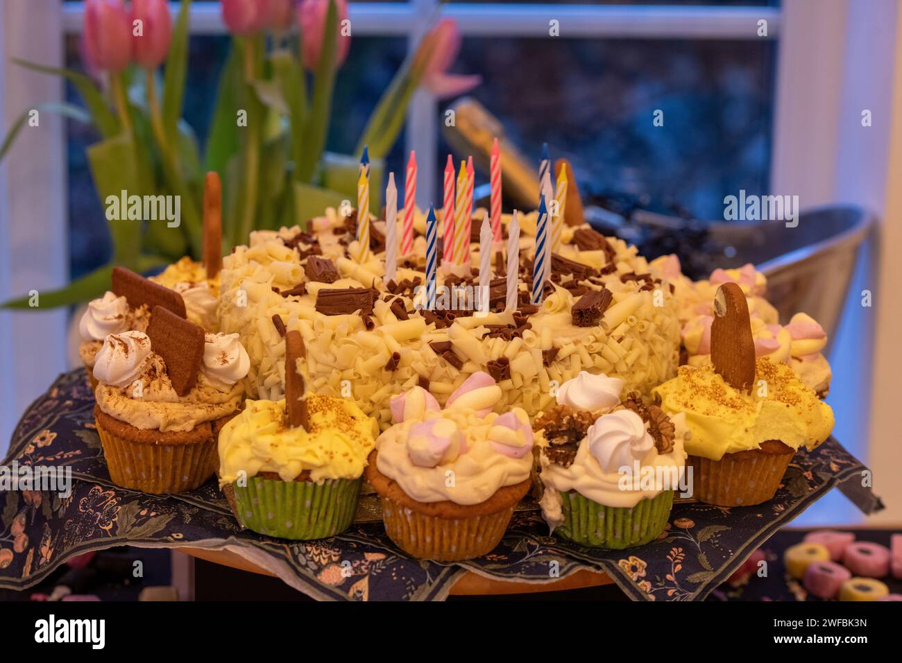 Una torta di compleanno al cioccolato bianco con candele spente circondata da cupcake fatti in casa coperti da decorazioni individuali e glassa di burro Foto Stock