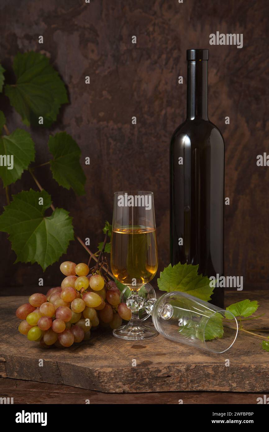 Il concetto di una deliziosa bevanda alcolica, vino e uva. Bottiglia di vino, bicchiere vuoto e bicchiere di vino bianco e mazzo di uva rosa su rustico scuro Foto Stock