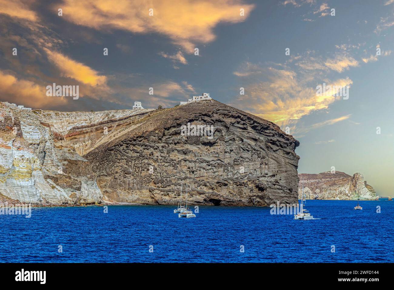 Vista panoramica della costa rocciosa vulcanica dell'isola di Santorini, con gli edifici della capitale Fira e le barche da diporto sul Mar Egeo. Luce del tramonto. Foto Stock