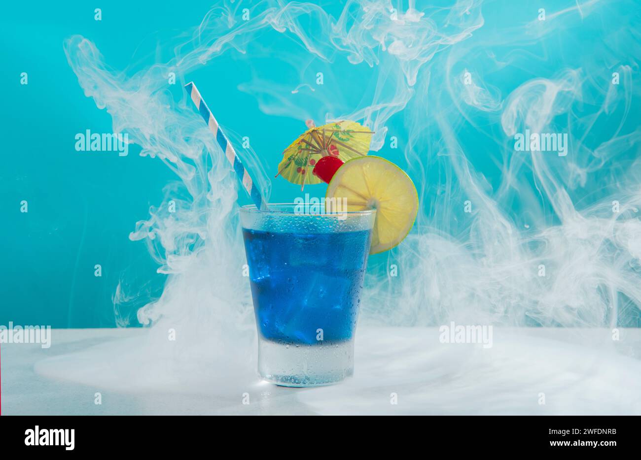 Cocktail dolce in un bicchiere di vetro freddo riempito di ghiaccio guarnito con fette di limone, ciliegia, ombrello giallo e sorbetto su un tavolo bianco contro bl leggero Foto Stock