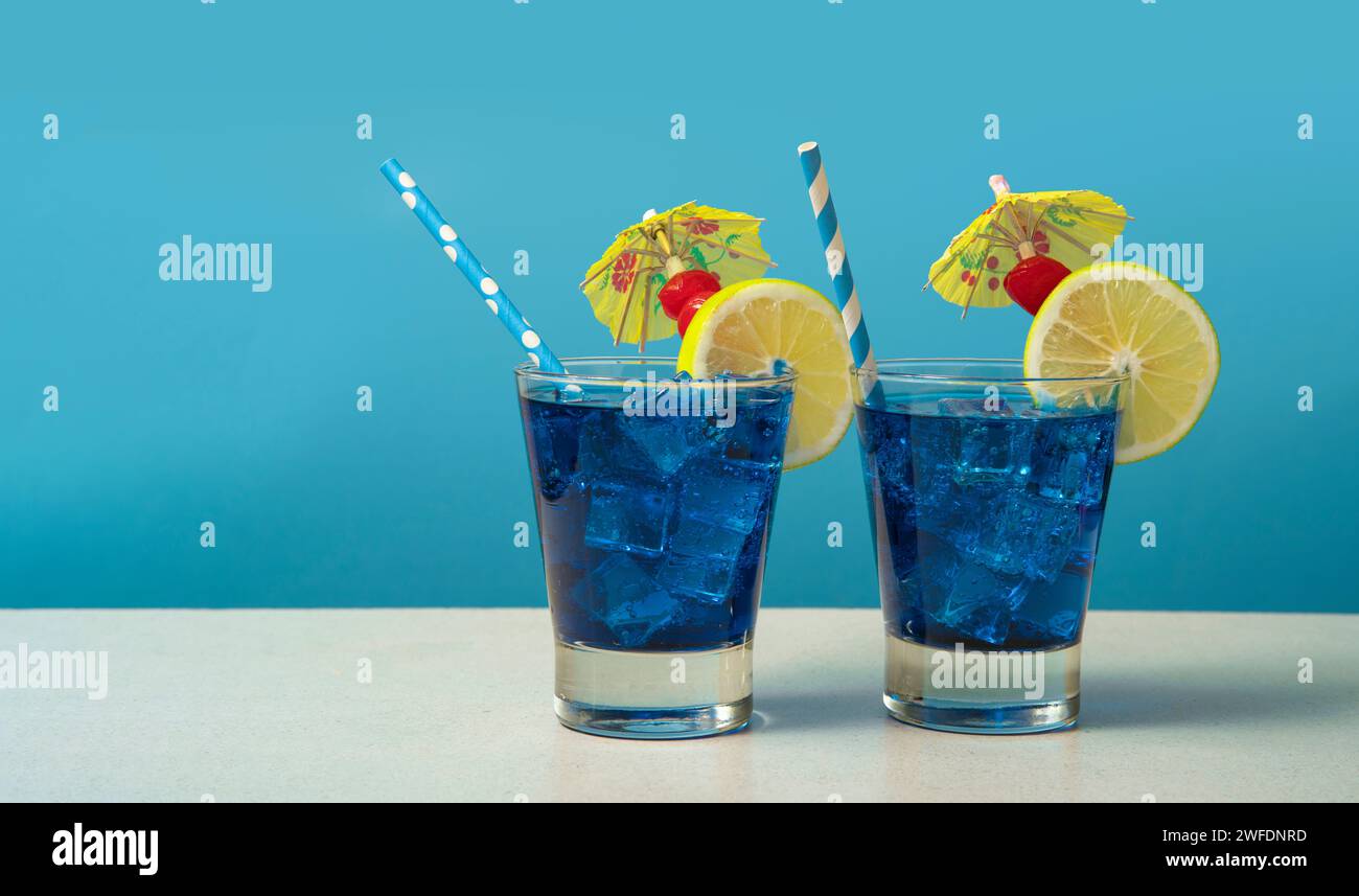 Due cocktail blu in un bicchiere pieno di ghiaccio guarnito con fette di limone, ciliegie, ombrelli gialli e sorbetti su un tavolo bianco contro una schiena blu Foto Stock