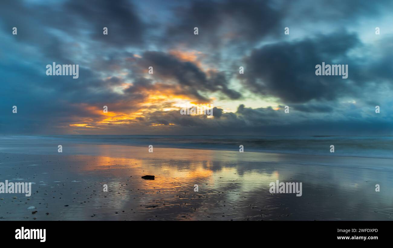 Nuvole minacciose e tramonto sul Mare del Nord lungo la costa dell'Olanda settentrionale. Le onde hanno lasciato un pezzo di legno sulla spiaggia. Foto Stock