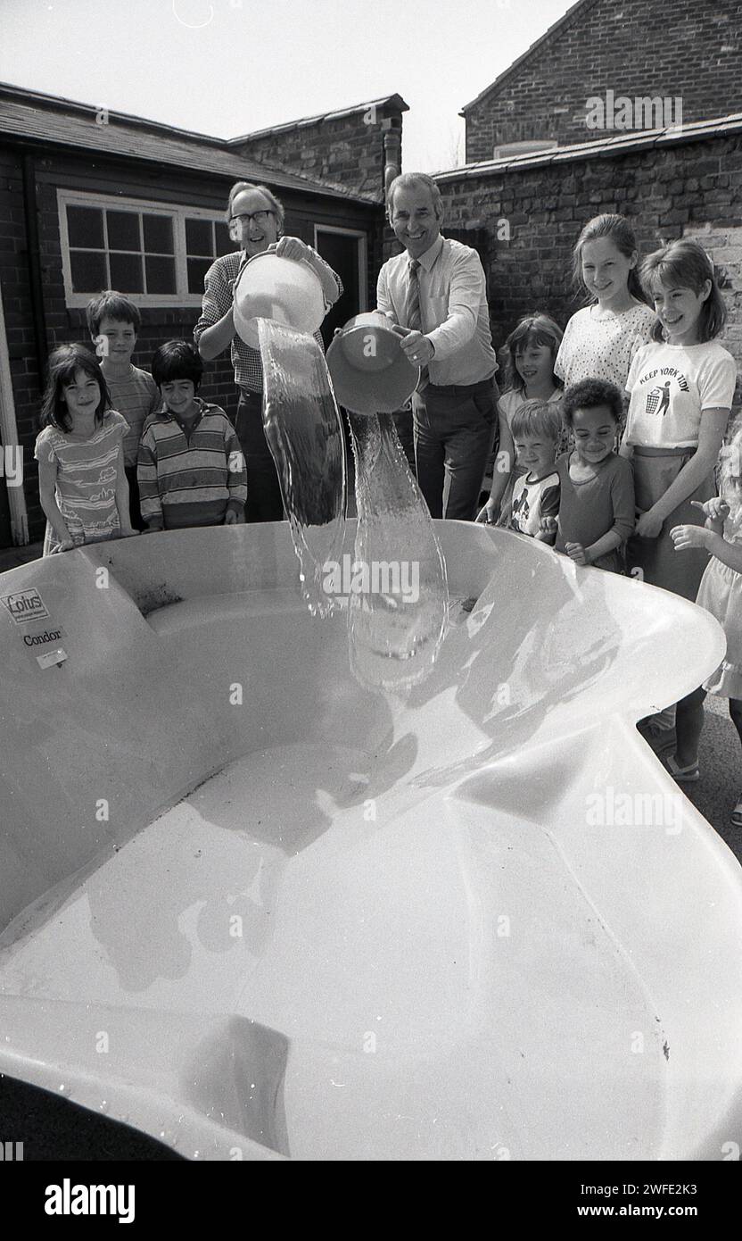 Anni '1980, battesimo del nuovo stagno scolastico, all'aperto in un parco giochi, studenti delle scuole che guardano insegnanti maschi versare acqua nel guscio di plastica, York, Inghilterra, Regno Unito. Foto Stock