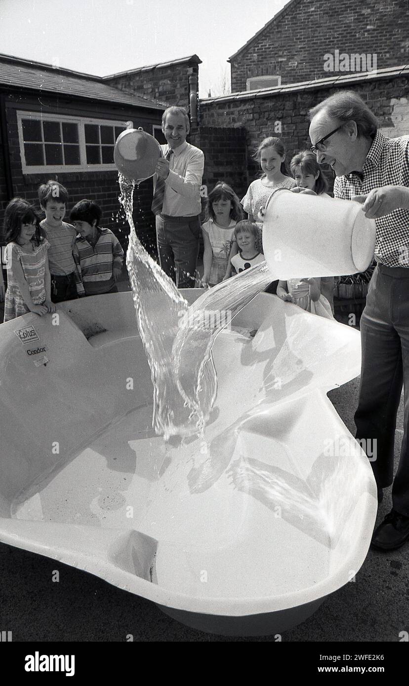 Anni '1980, battesimo del nuovo stagno scolastico, all'aperto in un parco giochi, studenti delle scuole che guardano insegnanti maschi versare acqua nel guscio di plastica, York, Inghilterra, Regno Unito. Foto Stock