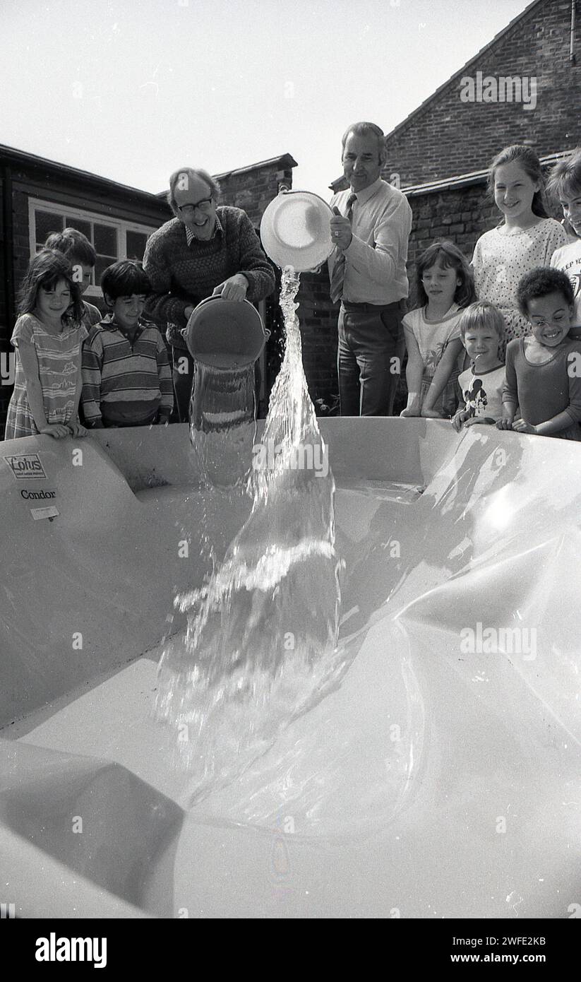 Anni '1980, battesimo del nuovo stagno scolastico, all'aperto in un parco giochi, studenti delle scuole che guardano insegnanti maschi versare acqua nel guscio di plastica, York, Inghilterra, Regno Unito. Foto Stock