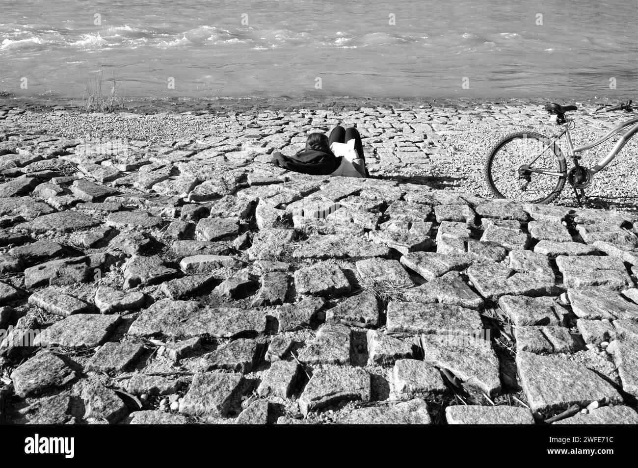 Donna che posa e legge un libro sulla riva del fiume e la sua bici sul lato Foto Stock