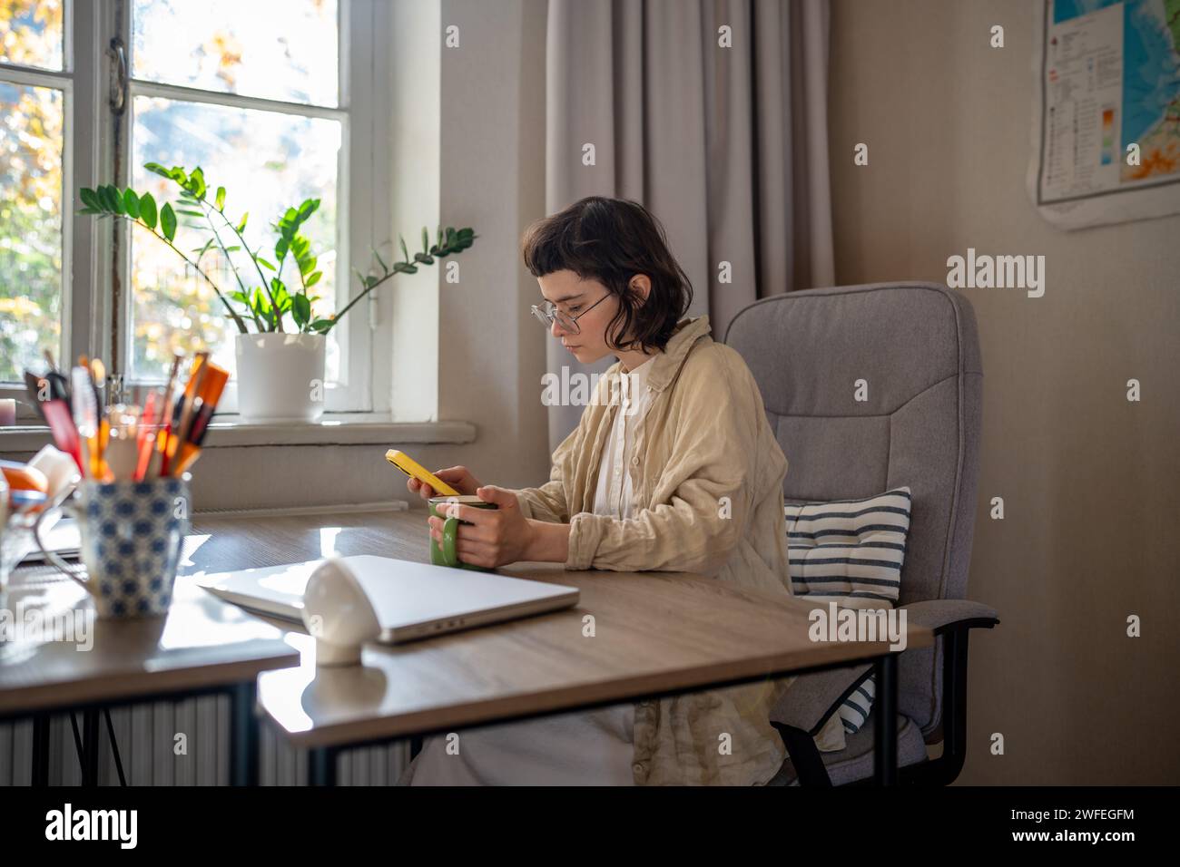 Teenager premuroso seduto al tavolo, che legge informazioni dal telefono cellulare, invia messaggi durante le pause di lavoro Foto Stock