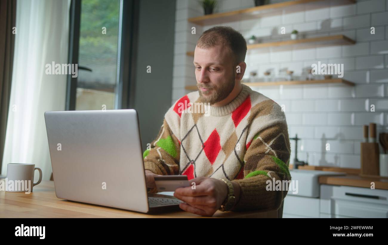 Uomo adulto seduto in cucina a casa, che inserisce il numero di carta di credito sul computer portatile per acquistare beni a distanza. La carta di credito è rifiutata o non utilizzata Foto Stock