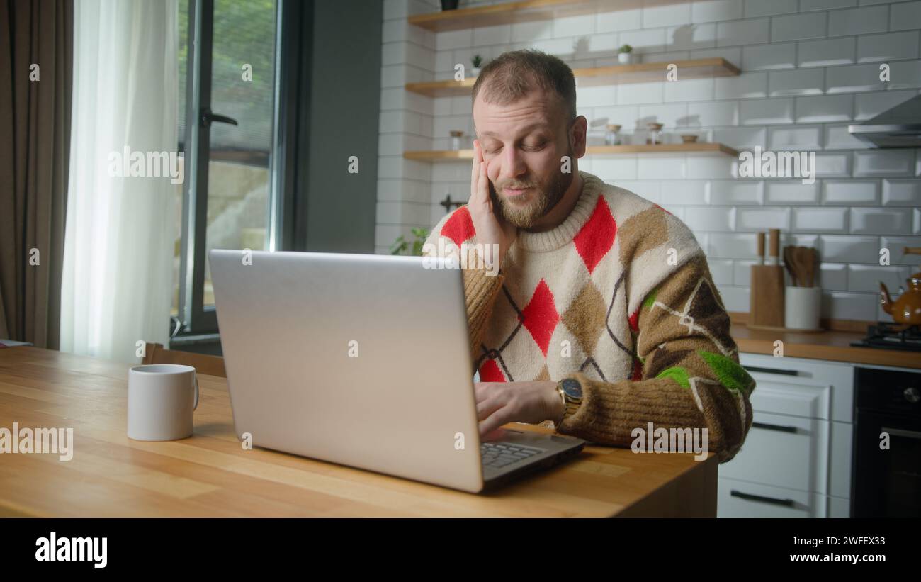 Un uomo adulto seduto in cucina a casa si strofina la testa mentre usa il computer portatile per lavorare. L'uomo stanco soffre di mal di testa o emicrania Foto Stock