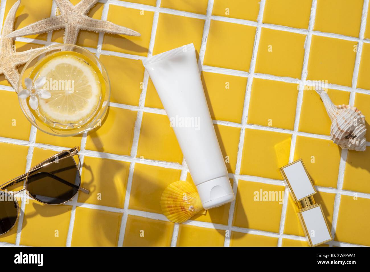 Stile di vita con occhiali da sole, rossetto, stelle marine, conchiglie decorate con un tubo senza etichetta su uno sfondo a mosaico giallo. Contenitore Foto Stock