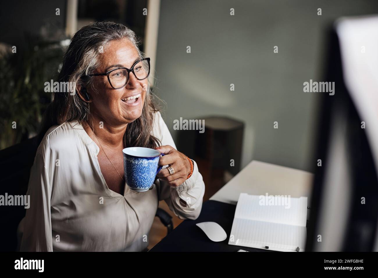 Donna d'affari matura che tiene cup mentre effettua videochiamate in ufficio Foto Stock