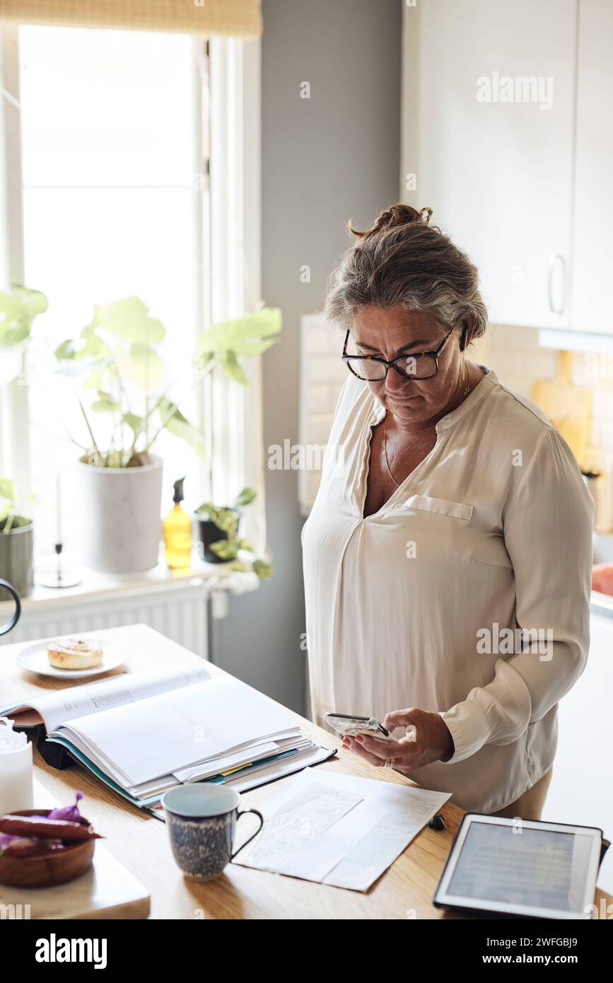 Donna matura che fotografa bollette finanziarie mentre è in cucina a casa Foto Stock
