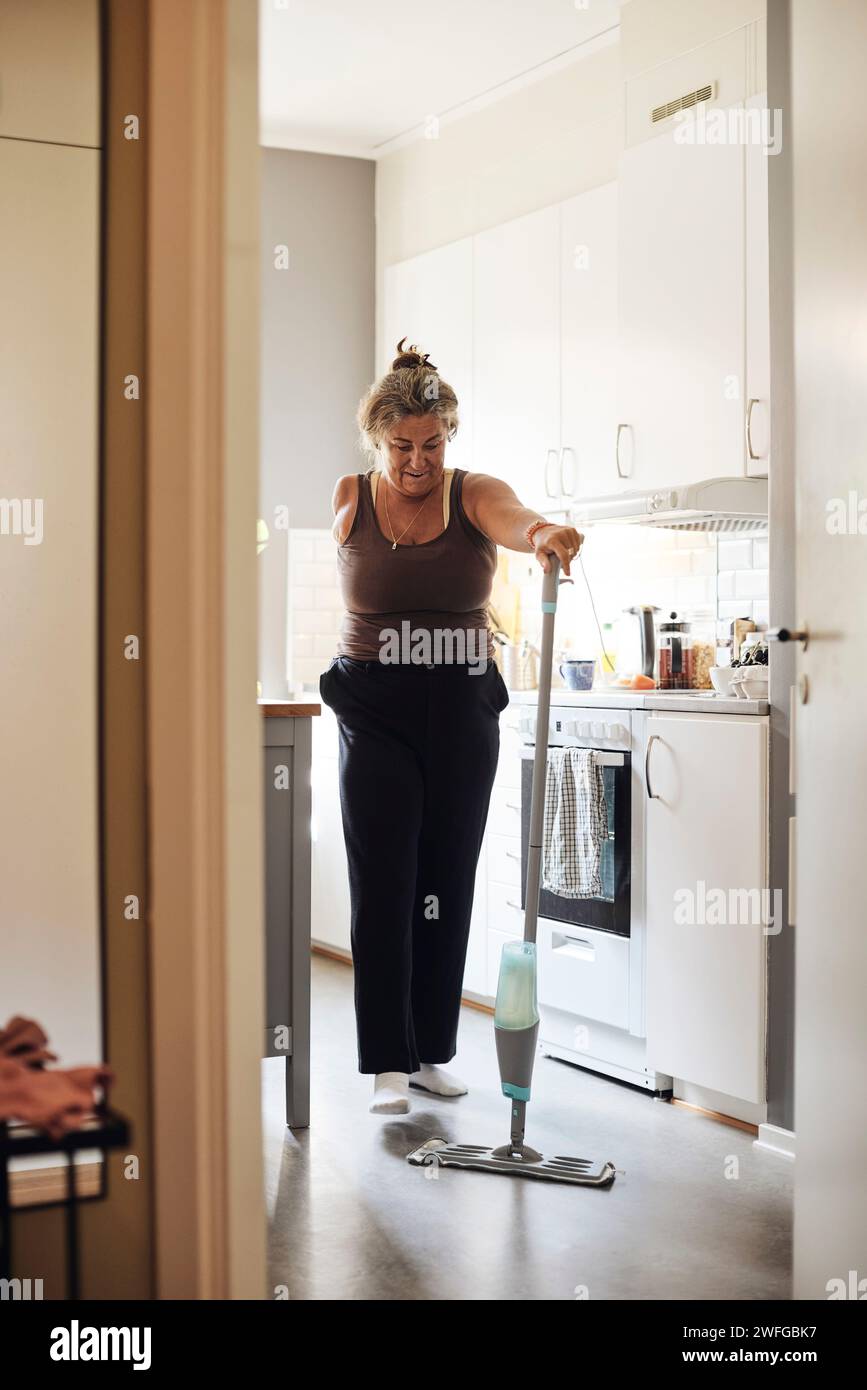 Donna matura con disabilità che pulisce il pavimento della cucina a casa Foto Stock