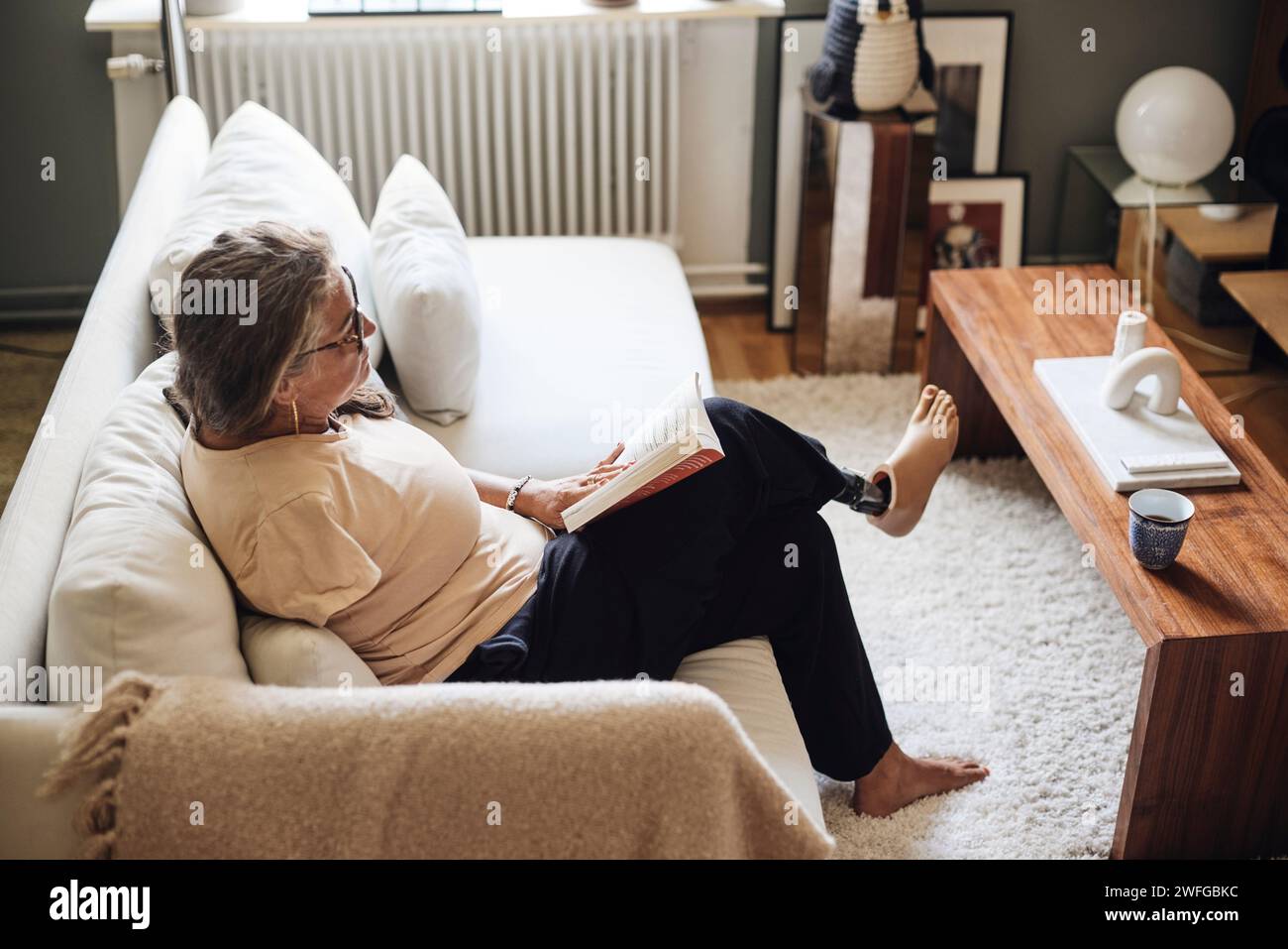 Vista ad alto angolo di una donna con disabilità che legge un libro mentre è seduta sul divano a casa Foto Stock