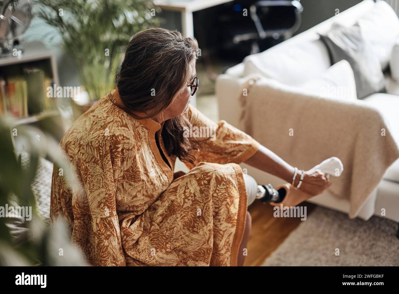 Donna matura con disabilità che indossa calzino su una gamba protesica mentre è seduta a casa Foto Stock