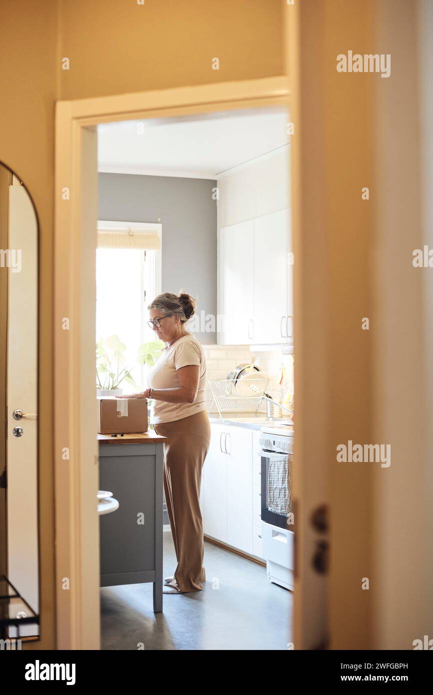 Vista laterale dell'intera lunghezza della donna matura che apre la confezione mentre è in cucina, vista dalla porta di casa Foto Stock