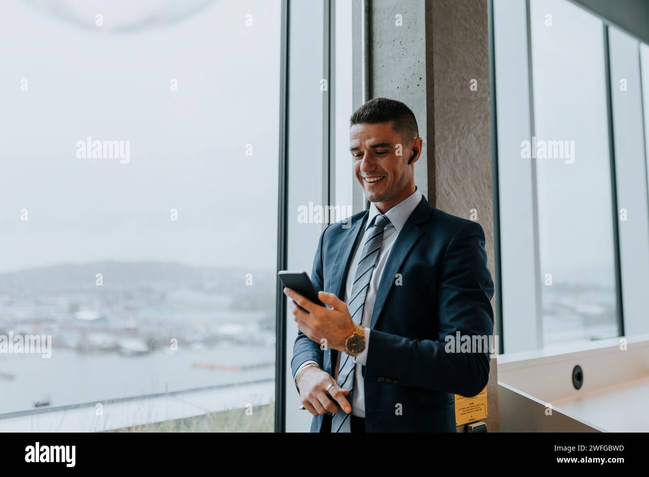 Giovane uomo d'affari sorridente con uno smartphone in piedi vicino alla finestra in ufficio Foto Stock