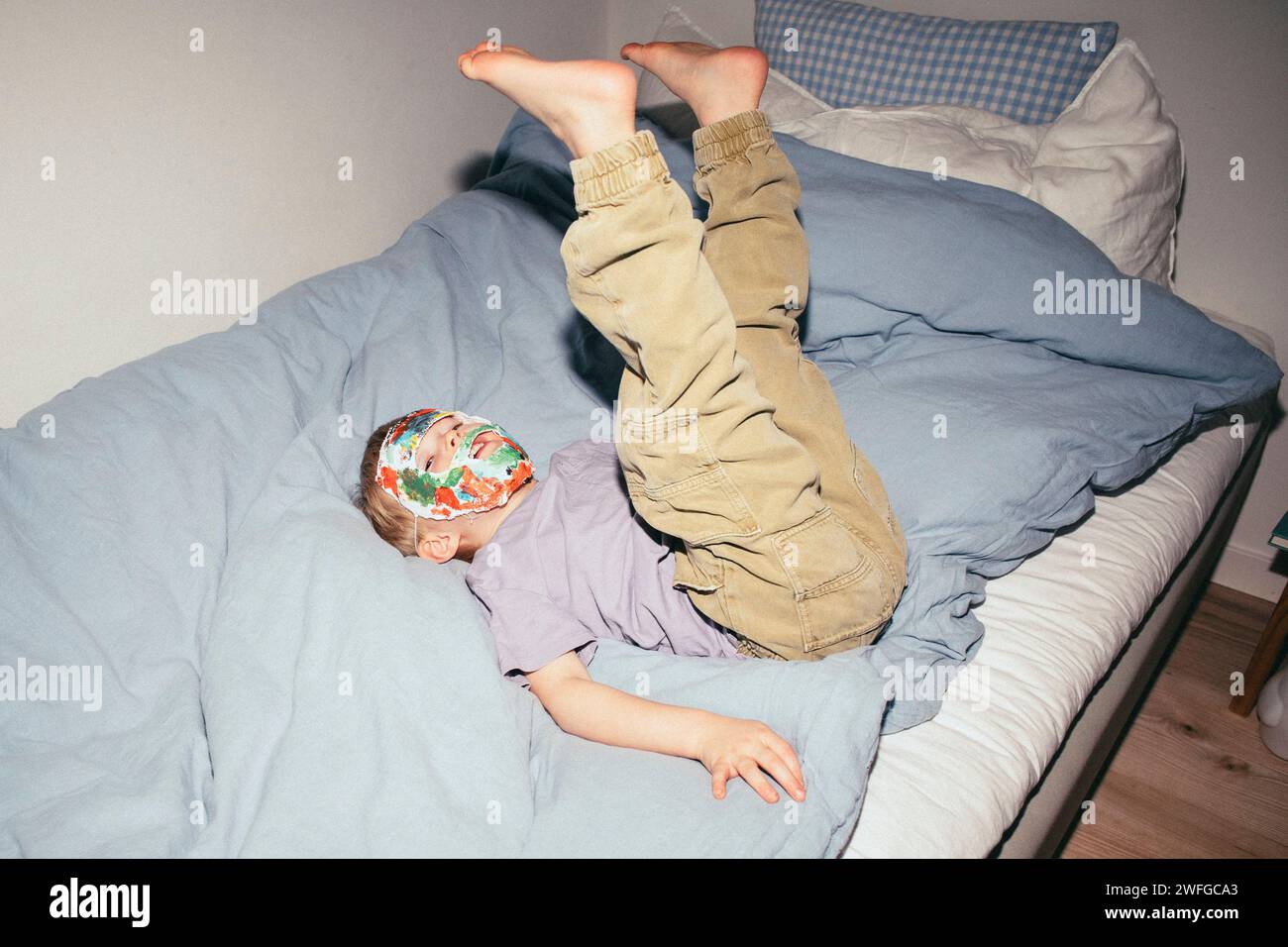 Ragazzo spensierato che indossa la maschera mentre è sdraiato sul letto con le gambe in alto a casa Foto Stock