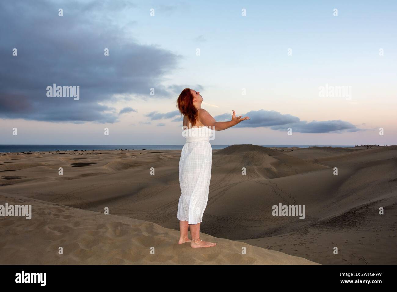 Una giovane ragazza si trova in un vestito bianco tra le dune di sabbia con il cielo e le nuvole nel tardo pomeriggio Foto Stock