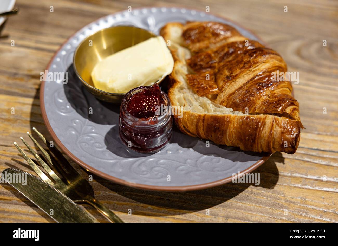 Croissant friabile servito con ricco burro e marmellata di fragole su un elegante piatto grigio, pronto per la colazione. Foto Stock