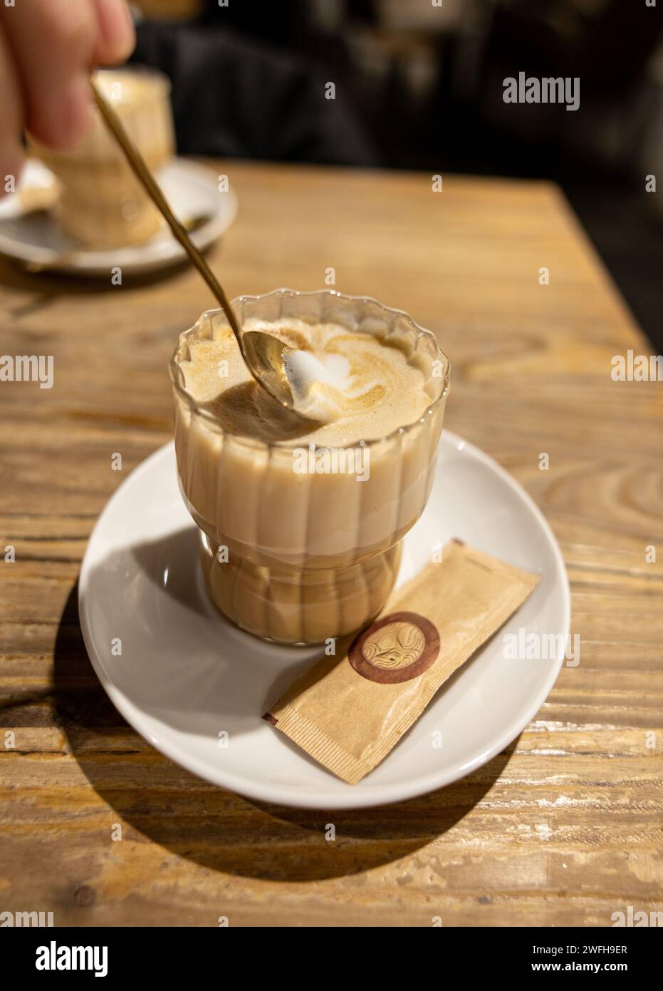 Gustando un cappuccino appena fatto, mescolando il latte con la schiuma in una tazza di vetro trasparente su un tavolo rustico in legno. Foto Stock