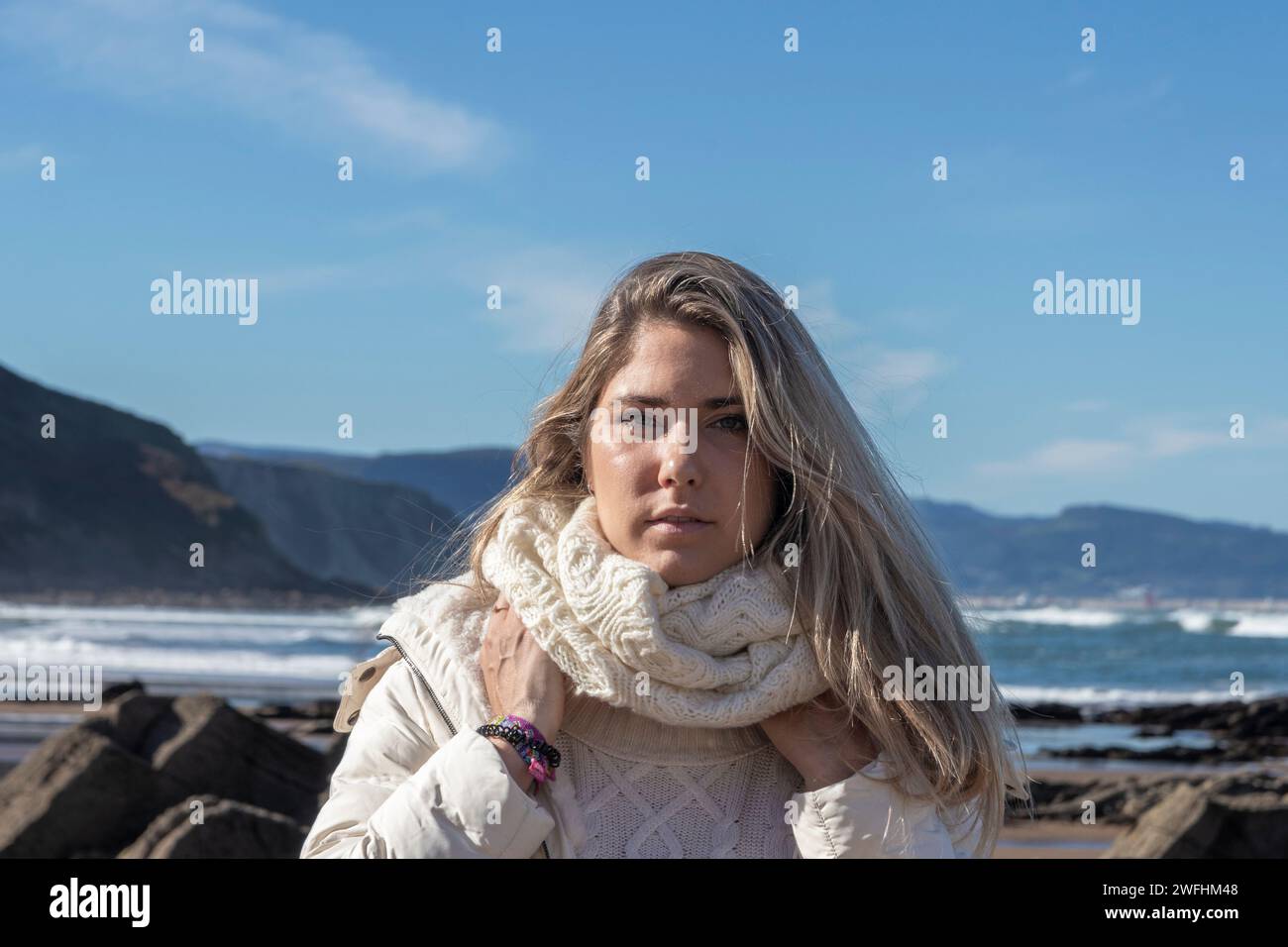donna in piedi su una costa rocciosa, che indossa un maglione e una giacca, con un oceano e scogliere sullo sfondo Foto Stock