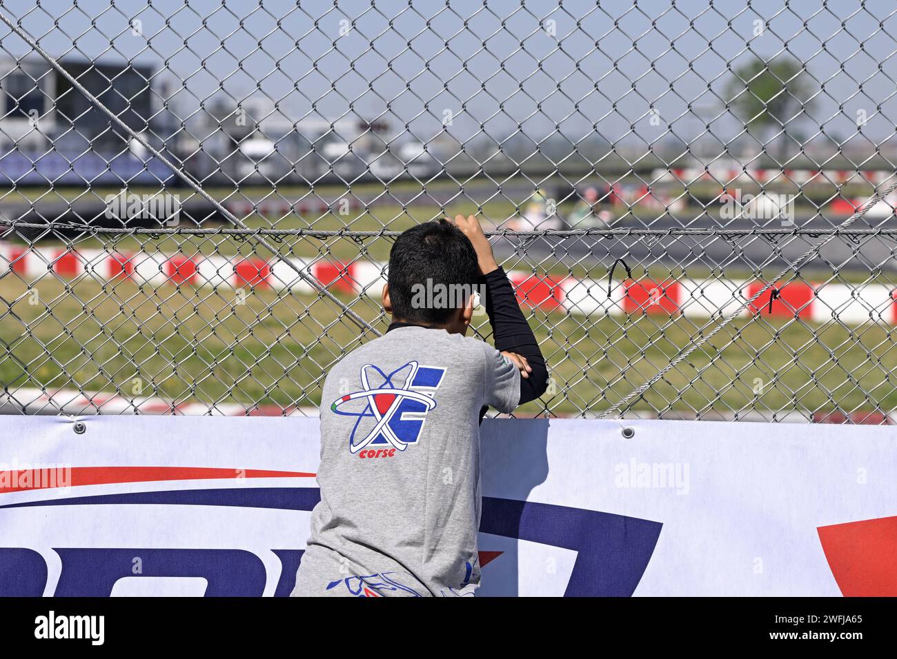 Giovane pilota appoggiato sulla rete e guardando le prove di gara sul circuito di go-kart Foto Stock