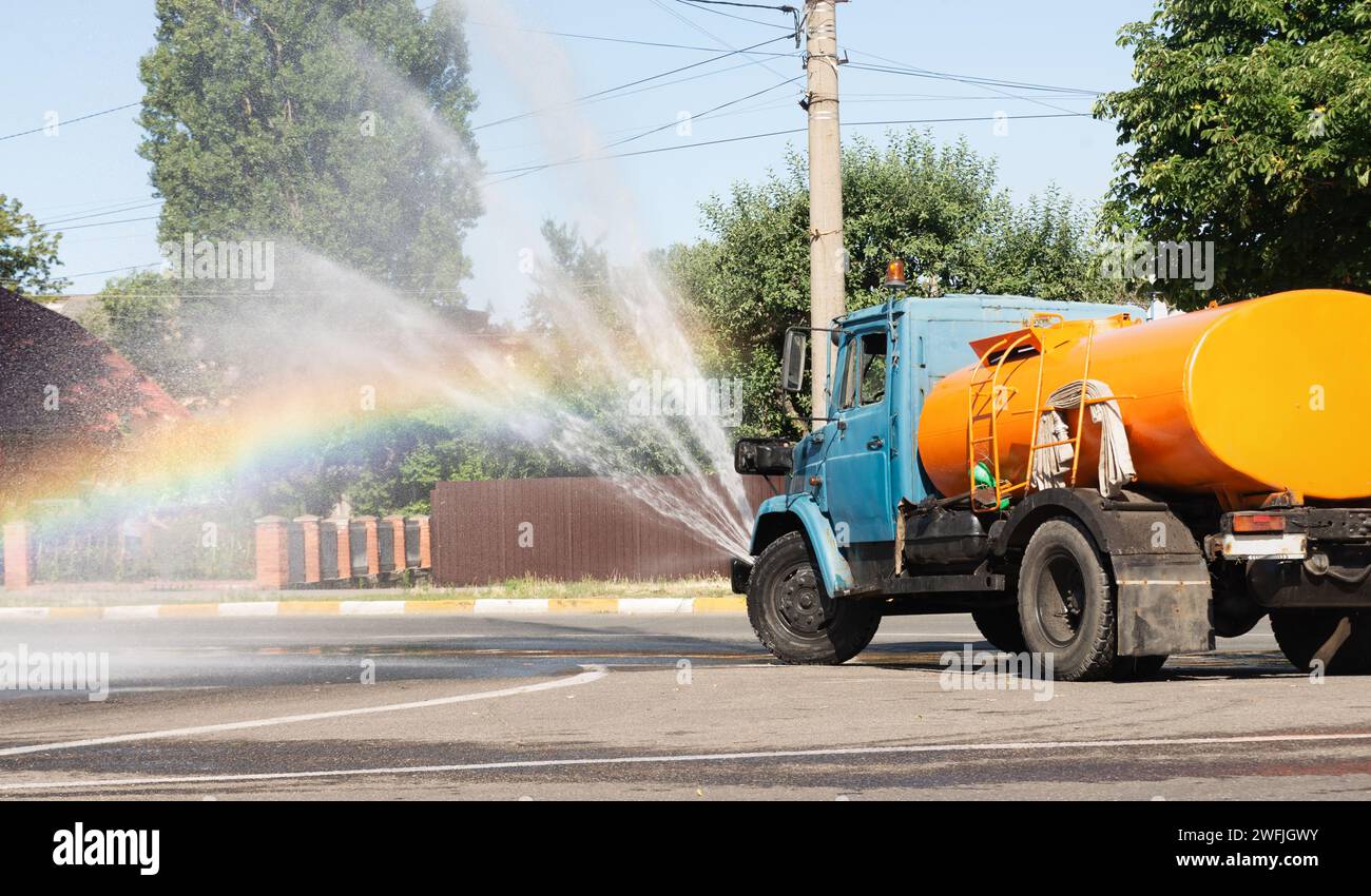 Il camion spruzza acqua sulla strada su uno sfondo arcobaleno. Servizi di assistenza in città. Foto Stock