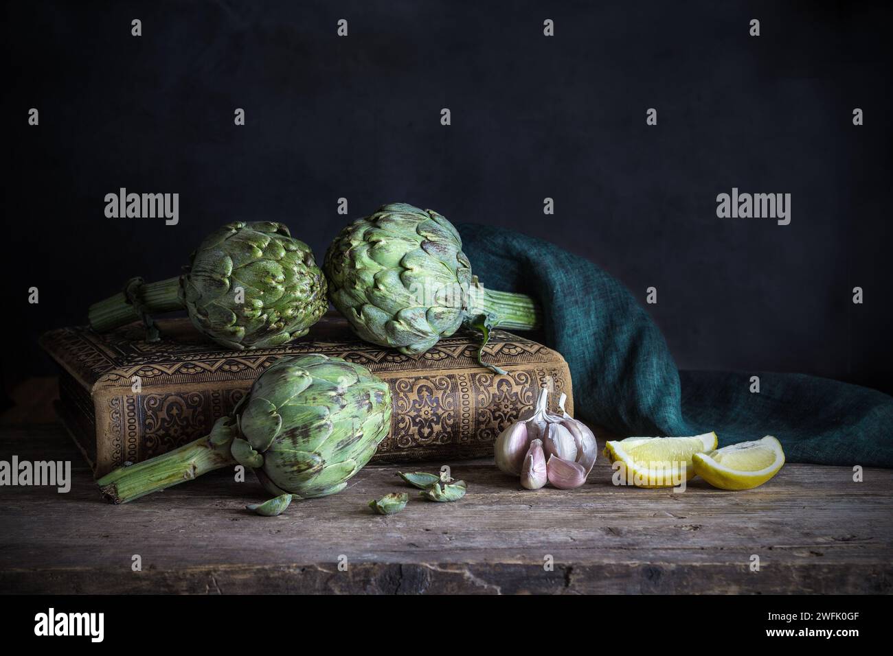 Carciofi freschi con aglio, limone su un libro antico e tovagliolo di lino - tonalità scure e lungose Foto Stock
