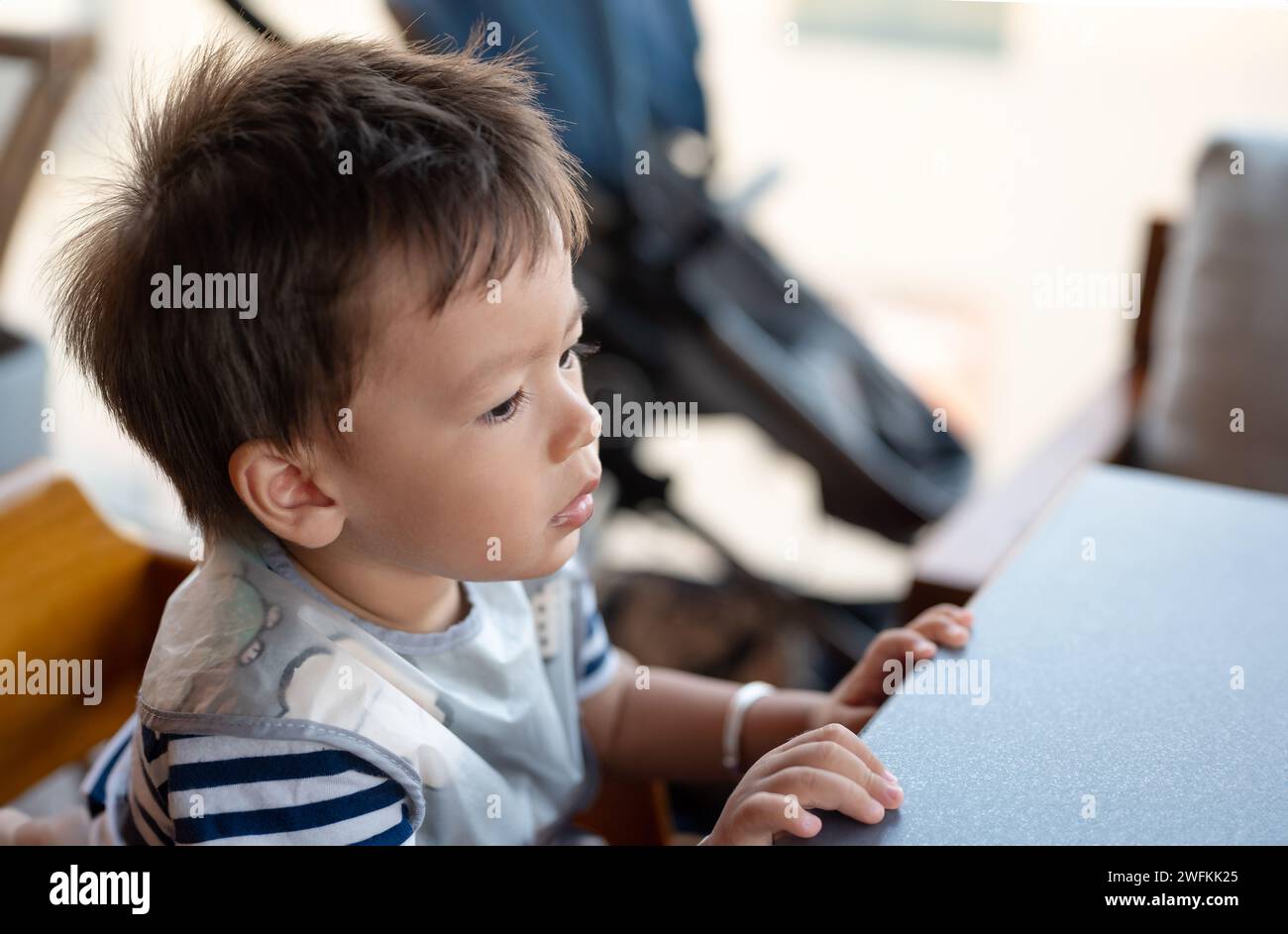 Ritratto ravvicinato di un piccolo e carino bambino vestito con una t-shirt a righe seduto su un seggiolone in un ristorante in attesa pazientemente del suo pranzo Foto Stock