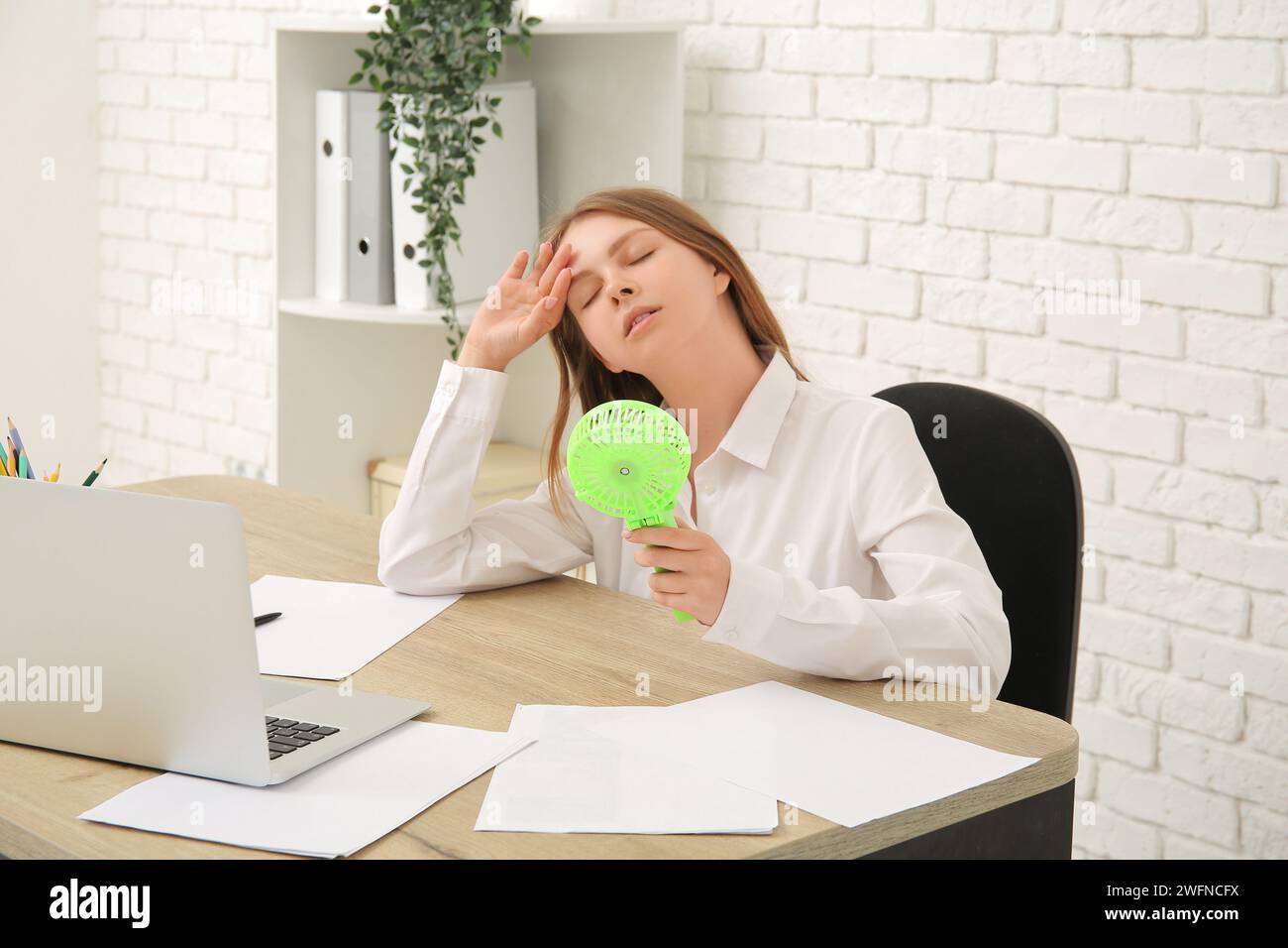 Bellissima giovane donna con mini ventilatore portatile in ufficio Foto Stock