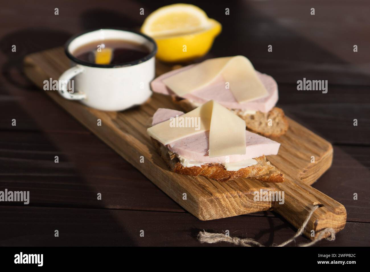 Panino con prosciutto, burro e formaggio a fette su un tagliere di legno vicino alla tazza di tè con limone Foto Stock