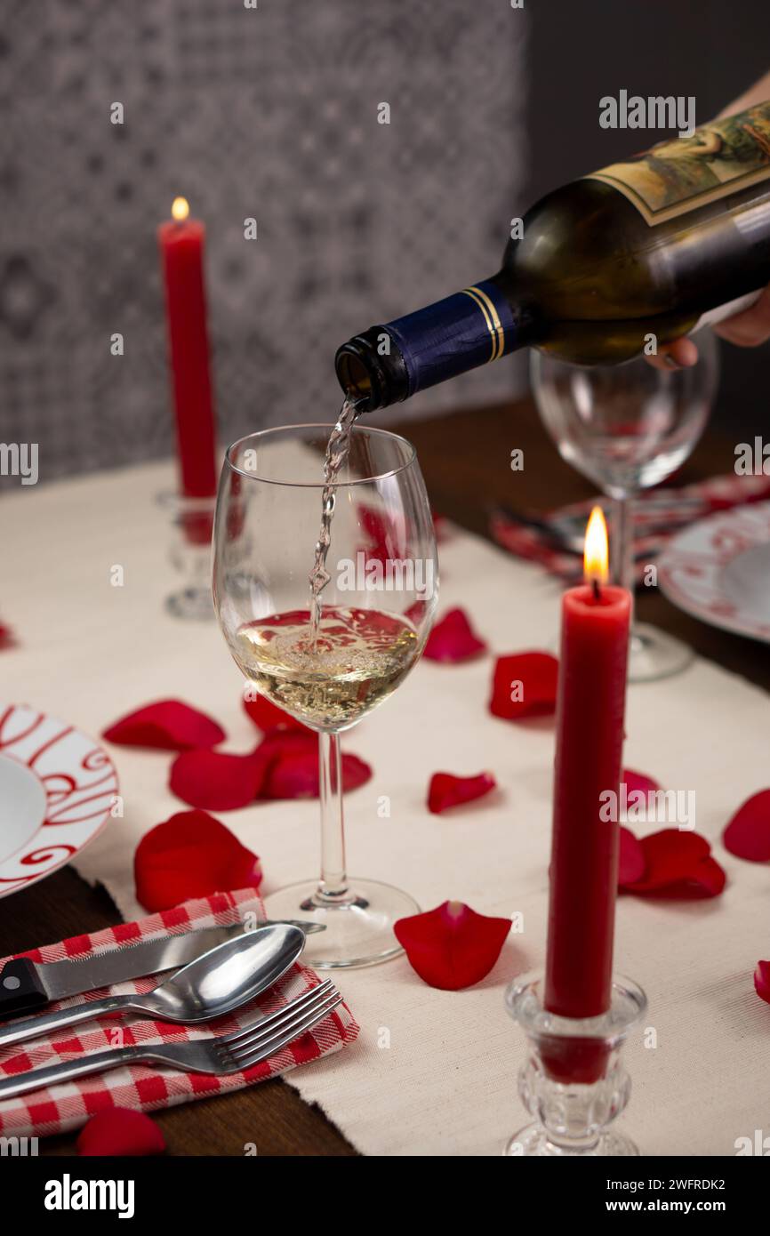 Serve a mano un bicchiere di vino bianco su un tavolo da pranzo per due con petali di rose rosse. C'è una candela rossa illuminata. Immagine verticale Foto Stock