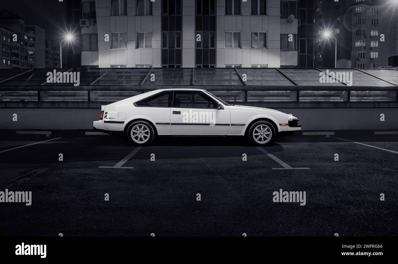 Coupé posteriore Toyota Celica Supra bianca parcheggiata sulla strada della città. Vista laterale dell'auto giapponese degli anni '1980 davanti allo sfondo urbano notturno. Foto Stock