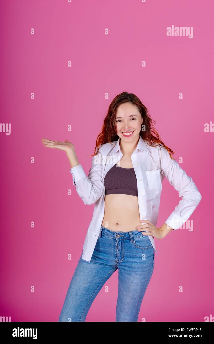 Felice donna rossa con una camicia bianca che si presenta con la mano, in piedi con sicurezza su uno sfondo rosa. Foto Stock